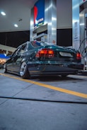 A dark-colored sedan is parked at a gas station during nighttime. The station has a modern design with bright lighting that reflects off the car’s polished surface. The car appears to have aftermarket wheels and lowered suspension. Fueling equipment and advertising signs are visible around the vehicle.