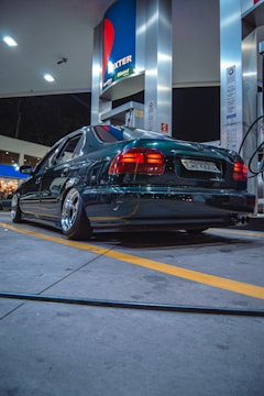 A dark-colored sedan is parked at a gas station during nighttime. The station has a modern design with bright lighting that reflects off the car’s polished surface. The car appears to have aftermarket wheels and lowered suspension. Fueling equipment and advertising signs are visible around the vehicle.