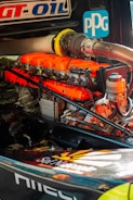 A detailed view of a high-performance engine with bright orange valve covers and various mechanical components. Logos and labels are visible on the engine and surrounding parts.
