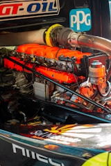 A detailed view of a high-performance engine with bright orange valve covers and various mechanical components. Logos and labels are visible on the engine and surrounding parts.