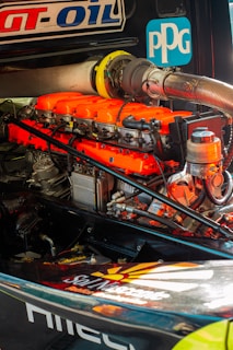 A detailed view of a high-performance engine with bright orange valve covers and various mechanical components. Logos and labels are visible on the engine and surrounding parts.