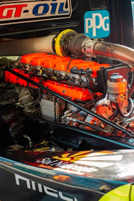 A detailed view of a high-performance engine with bright orange valve covers and various mechanical components. Logos and labels are visible on the engine and surrounding parts.