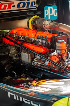 A detailed view of a high-performance engine with bright orange valve covers and various mechanical components. Logos and labels are visible on the engine and surrounding parts.