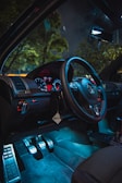 A car interior with a modern dashboard featuring illuminated gauges and a steering wheel. The ambient lighting creates a stylish, futuristic atmosphere, highlighting the pedals and upholstery. The window view includes blurred trees and a dark sky, suggesting nighttime from inside a parked car.