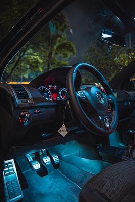 A car interior with a modern dashboard featuring illuminated gauges and a steering wheel. The ambient lighting creates a stylish, futuristic atmosphere, highlighting the pedals and upholstery. The window view includes blurred trees and a dark sky, suggesting nighttime from inside a parked car.