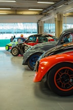 Several vintage racing cars are lined up inside a garage. The cars feature vibrant color schemes and are equipped with racing modifications. The setting is a spacious, well-lit area with a view of the race track in the background.