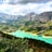 A stunning landscape featuring a bright turquoise river winding through lush green forests. The scene is set against a backdrop of rocky, towering mountains under a partly cloudy sky.