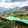 A stunning landscape featuring a bright turquoise river winding through lush green forests. The scene is set against a backdrop of rocky, towering mountains under a partly cloudy sky.