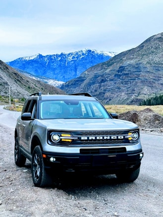 A rugged SUV is parked on a gravel road against the backdrop of towering mountains with snow-capped peaks. The sky is overcast, and the vehicle's headlights are on, suggesting a chilly or mountainous setting.