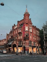 A historic red brick building with a turret, featuring a neon sign for a hotel. The architecture includes arched windows and decorative stonework. People are walking and cycling in front of the building on a street with tram lines overhead. Trees are visible to the side, contributing to an urban atmosphere.