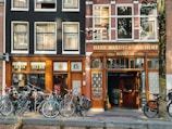A street scene featuring two adjacent storefronts with bicycles parked in front. The left storefront displays the sign 'Sensi Seeds' with a classic black and wood facade, while the right is the 'Hash Marihuana & Hemp Museum' with similar architectural styling. Large windows with curtains line the upper floors, and a tree is visible on the right.