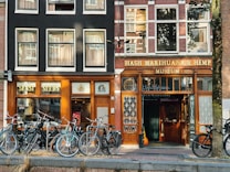 A street scene featuring two adjacent storefronts with bicycles parked in front. The left storefront displays the sign 'Sensi Seeds' with a classic black and wood facade, while the right is the 'Hash Marihuana & Hemp Museum' with similar architectural styling. Large windows with curtains line the upper floors, and a tree is visible on the right.