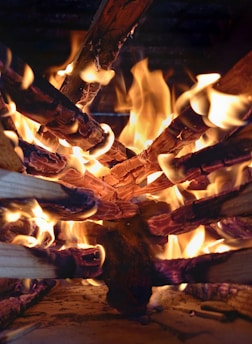 A close-up view of burning logs arranged in a circular pattern with bright, orange flames and charring visible on the wood surfaces.