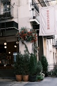 A quaint sidewalk scene featuring a restaurant entrance with lush greenery in large pots and a vibrant display of red and orange flowers climbing the wall. The building has a rustic charm with a visible sign that reads 'Boucherie' and decorative railings. Inside, there is a cozy bar area with hanging lights and a warm ambience.