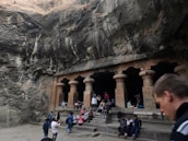 A group of people are gathered in front of ancient stone structures carved into a rocky landscape. The architecture features large pillars and an open facade, set against a backdrop of rugged, natural rock formations. The scene suggests a historical or tourist site with visitors resting, taking photos, and exploring the surroundings.
