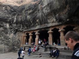 A group of people are gathered in front of ancient stone structures carved into a rocky landscape. The architecture features large pillars and an open facade, set against a backdrop of rugged, natural rock formations. The scene suggests a historical or tourist site with visitors resting, taking photos, and exploring the surroundings.