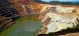 A large open-pit mine with terraced layers and a water body at the bottom. The exposed earth shows a variety of colors, indicating different soil and rock types. There is construction equipment visible on the terraces and some vegetation in the surrounding area.