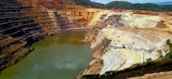 A large open-pit mine with terraced layers and a water body at the bottom. The exposed earth shows a variety of colors, indicating different soil and rock types. There is construction equipment visible on the terraces and some vegetation in the surrounding area.