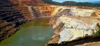 A large open-pit mine with terraced layers and a water body at the bottom. The exposed earth shows a variety of colors, indicating different soil and rock types. There is construction equipment visible on the terraces and some vegetation in the surrounding area.