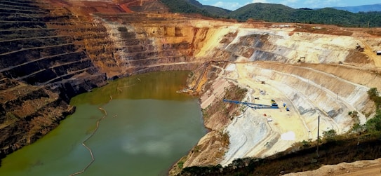 A large open-pit mine with terraced layers and a water body at the bottom. The exposed earth shows a variety of colors, indicating different soil and rock types. There is construction equipment visible on the terraces and some vegetation in the surrounding area.