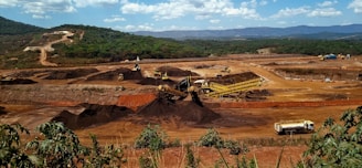 A large mining site is spread across a vast landscape with multiple heavy machinery and trucks in operation. The site has a variety of soil and rock formations, with visible conveyor belts and excavators working. The background features green hills and a partly cloudy sky.