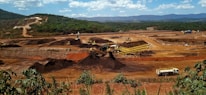 A large mining site is spread across a vast landscape with multiple heavy machinery and trucks in operation. The site has a variety of soil and rock formations, with visible conveyor belts and excavators working. The background features green hills and a partly cloudy sky.