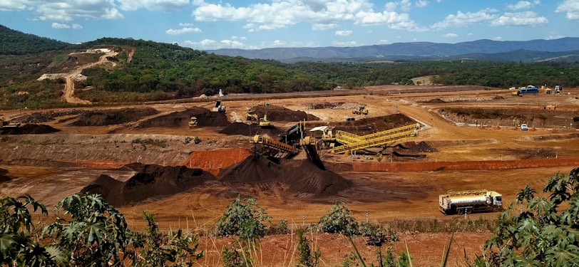 A large mining site is spread across a vast landscape with multiple heavy machinery and trucks in operation. The site has a variety of soil and rock formations, with visible conveyor belts and excavators working. The background features green hills and a partly cloudy sky.