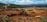 A large mining site is spread across a vast landscape with multiple heavy machinery and trucks in operation. The site has a variety of soil and rock formations, with visible conveyor belts and excavators working. The background features green hills and a partly cloudy sky.
