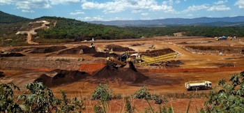 A large mining site is spread across a vast landscape with multiple heavy machinery and trucks in operation. The site has a variety of soil and rock formations, with visible conveyor belts and excavators working. The background features green hills and a partly cloudy sky.