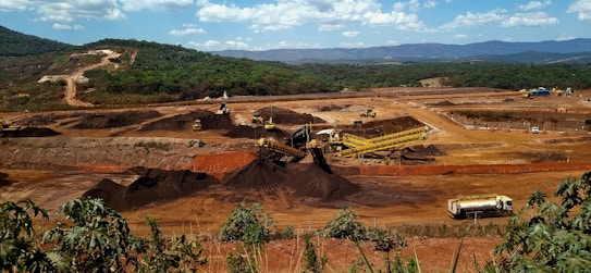 A large mining site is spread across a vast landscape with multiple heavy machinery and trucks in operation. The site has a variety of soil and rock formations, with visible conveyor belts and excavators working. The background features green hills and a partly cloudy sky.