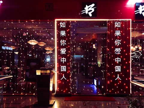 The entrance to a restaurant is adorned with vertical string lights, creating a warm and inviting glow. The signage in Chinese characters is prominently displayed on illuminated panels. The interior is visible through glass, showcasing hanging pendant lights and neatly arranged tables.