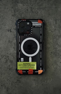 A smartphone with a black and transparent case featuring the brand name 'Casetify'. The case has a prominent white circular design labeled 'Duty Free Goods'. There is a bright yellow label that reads 'Warning! Duty Free Purchase' and several red tamper-evident stickers affixed to it. The phone is placed on a textured gray surface.