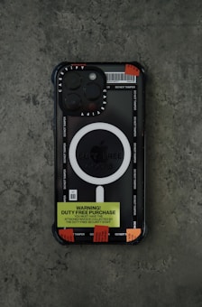 A smartphone with a black and transparent case featuring the brand name 'Casetify'. The case has a prominent white circular design labeled 'Duty Free Goods'. There is a bright yellow label that reads 'Warning! Duty Free Purchase' and several red tamper-evident stickers affixed to it. The phone is placed on a textured gray surface.