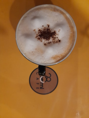 A top view of a cappuccino cup with frothy milk and a sprinkle of cocoa powder in the center. The surface beneath the cup is a warm yellow, and a logo for '28ml Café' is visible.