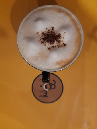 A top view of a cappuccino cup with frothy milk and a sprinkle of cocoa powder in the center. The surface beneath the cup is a warm yellow, and a logo for '28ml Caf&eacute;' is visible.