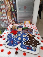 A large, creatively decorated cake featuring various thematic sections and small decorative items such as candles, flowers, and miniatures of plants and animals. The cake has segments in different colors, including blue, red, brown, and white, and is bordered with white icing. It is surrounded by scattered red petals and small pinecones.