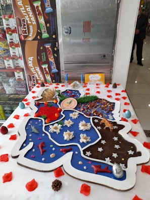 A large, creatively decorated cake featuring various thematic sections and small decorative items such as candles, flowers, and miniatures of plants and animals. The cake has segments in different colors, including blue, red, brown, and white, and is bordered with white icing. It is surrounded by scattered red petals and small pinecones.