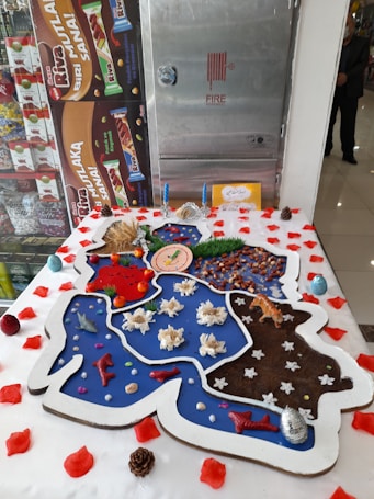 A large, creatively decorated cake featuring various thematic sections and small decorative items such as candles, flowers, and miniatures of plants and animals. The cake has segments in different colors, including blue, red, brown, and white, and is bordered with white icing. It is surrounded by scattered red petals and small pinecones.