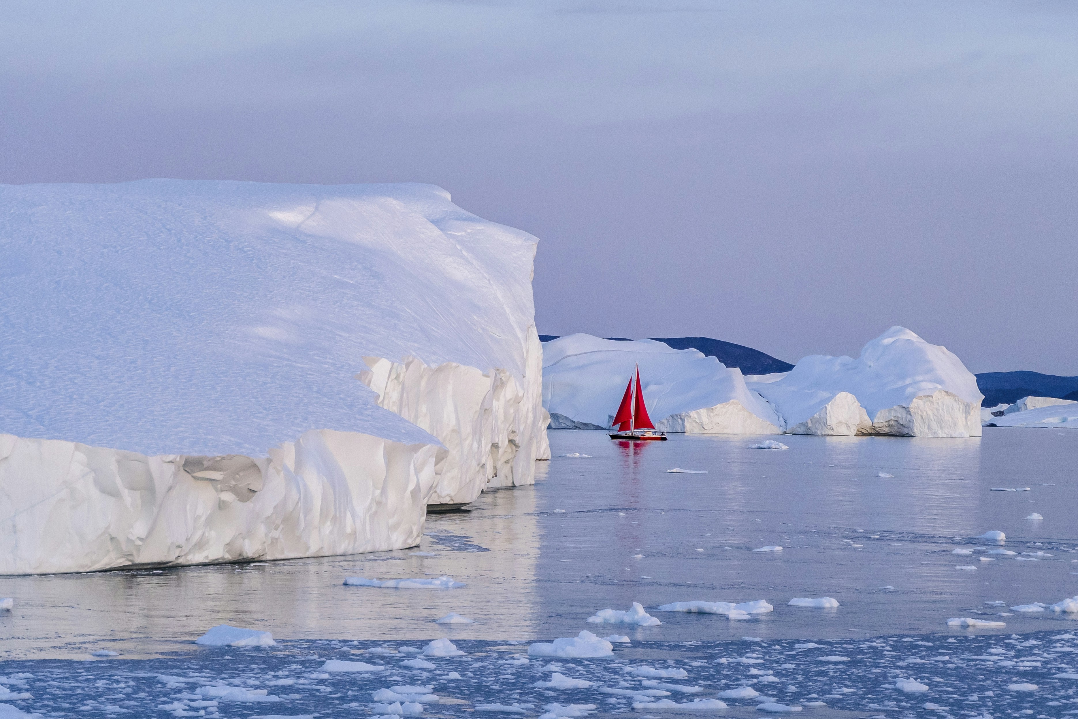 a red sailboat in the water near icebergs, A boat with red sails stands out against a back drop of giant icebergs.