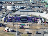 Aerial view of a large outdoor stadium setup for a concert or event, featuring a stage with vibrant purple and green decorations and the word 'GLUCKSOEFUHLE' displayed prominently. Surrounding the stage are various technical equipment and structures, including lighting and sound rigs. The seating areas in the stadium appear to be empty. Around the stadium, there are numerous temporary structures, vehicles, and equipment, indicating preparations for the event.