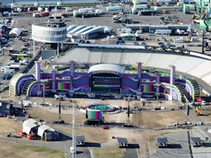 Aerial view of a large outdoor stadium setup for a concert or event, featuring a stage with vibrant purple and green decorations and the word 'GLUCKSOEFUHLE' displayed prominently. Surrounding the stage are various technical equipment and structures, including lighting and sound rigs. The seating areas in the stadium appear to be empty. Around the stadium, there are numerous temporary structures, vehicles, and equipment, indicating preparations for the event.
