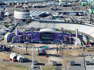 Aerial view of a large outdoor stadium setup for a concert or event, featuring a stage with vibrant purple and green decorations and the word 'GLUCKSOEFUHLE' displayed prominently. Surrounding the stage are various technical equipment and structures, including lighting and sound rigs. The seating areas in the stadium appear to be empty. Around the stadium, there are numerous temporary structures, vehicles, and equipment, indicating preparations for the event.