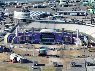 Aerial view of a large outdoor stadium setup for a concert or event, featuring a stage with vibrant purple and green decorations and the word 'GLUCKSOEFUHLE' displayed prominently. Surrounding the stage are various technical equipment and structures, including lighting and sound rigs. The seating areas in the stadium appear to be empty. Around the stadium, there are numerous temporary structures, vehicles, and equipment, indicating preparations for the event.