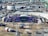 Aerial view of a large outdoor stadium setup for a concert or event, featuring a stage with vibrant purple and green decorations and the word 'GLUCKSOEFUHLE' displayed prominently. Surrounding the stage are various technical equipment and structures, including lighting and sound rigs. The seating areas in the stadium appear to be empty. Around the stadium, there are numerous temporary structures, vehicles, and equipment, indicating preparations for the event.