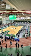 A large indoor arena with a basketball court at the center, surrounded by multiple stalls set up for a graduate and professional schools fair. The area is bustling with people walking around, interacting, and gathering information at various booths. A prominent digital screen displays information about the event. Overhead, banners are visible, indicating past sports events.