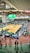 A large indoor arena with a basketball court at the center, surrounded by multiple stalls set up for a graduate and professional schools fair. The area is bustling with people walking around, interacting, and gathering information at various booths. A prominent digital screen displays information about the event. Overhead, banners are visible, indicating past sports events.