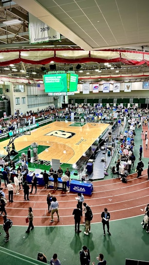 A large indoor arena with a basketball court at the center, surrounded by multiple stalls set up for a graduate and professional schools fair. The area is bustling with people walking around, interacting, and gathering information at various booths. A prominent digital screen displays information about the event. Overhead, banners are visible, indicating past sports events.