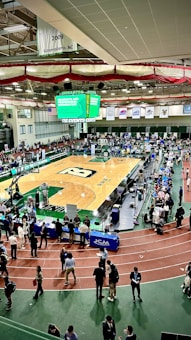 A large indoor arena with a basketball court at the center, surrounded by multiple stalls set up for a graduate and professional schools fair. The area is bustling with people walking around, interacting, and gathering information at various booths. A prominent digital screen displays information about the event. Overhead, banners are visible, indicating past sports events.