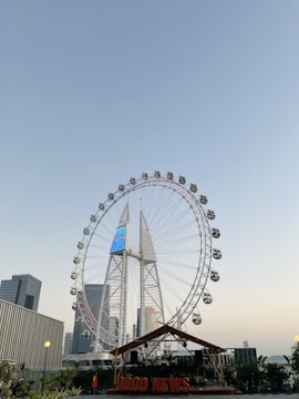 A large Ferris wheel stands prominently against a clear blue sky, surrounded by modern skyscrapers. Below the Ferris wheel, there is a structure with an orange sign reading 'GOOD NEWS', accented by greenery and a lit lamp. The sun is setting, casting a warm glow over the scene.