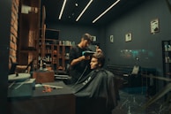 A modern barbershop with a stylish interior featuring exposed brick walls, sleek shelving filled with products, and framed photos on the walls. A barber is attentively working on a customer's haircut, using a hairdryer. The atmosphere is cozy and well-organized.